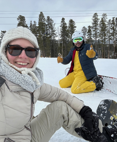 Two Principle employees sitting on a snowy hill with cedar trees in the background, wearing snowboard outfits with snowboards. Both are smiling, and one is giving two thumbs-up.